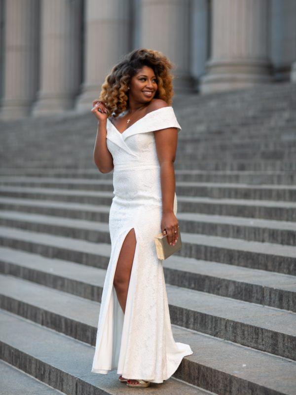 African American woman in a long white off-the-shoulder dress and gold accessories standing on marble steps outside a grand building