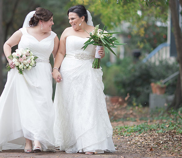 Two brides holding hands and their bridal bouquets while walking through a park